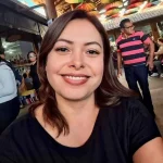 Software: Uma mulher com longos cabelos castanhos sorri para a câmera, vestindo uma camisa preta. Ao fundo, pessoas passeiam por um mercado aberto com guarda-chuvas coloridos pendurados acima delas, lembrando uma vibrante loja de software movimentada com atividade e oferecendo entrega imediata.