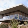 Software: Uma casa moderna em balanço com janelas de vidro expansivas se estende sobre uma paisagem rochosa, espelhando o design elegante do Lumion 2023 – Renderização 3D em Tempo Real para Arquitetura. A estrutura angular, completa com um telhado plano, está aninhada entre arbustos e pedras sob um céu parcialmente nublado.