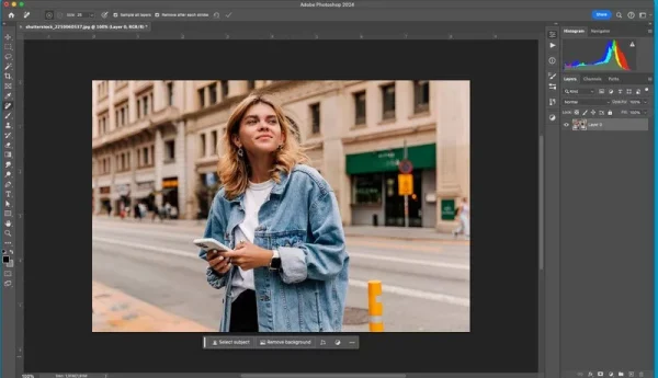 Software: Uma pessoa está em uma rua da cidade, segurando um telefone e vestindo uma jaqueta jeans, aparentemente olhando para a distância. A imagem está aberta no Adobe Photoshop 2024 - Completo com Licença em uma tela de computador, possivelmente após uma entrega instantânea de software.