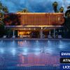 Software: Uma casa contemporânea e iluminada com uma piscina ampla na frente é envolvida por vegetação verdejante. O céu está parcialmente nublado, contribuindo para um ambiente tranquilo à noite. No canto inferior direito, há três caixas de texto com palavras em português, que lembram a navegação de software ao comprar um Lumion 12 - Licença Vitalícia online.