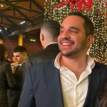 Software: Um homem de terno escuro e camisa branca de colarinho aberto sorri enquanto está em um evento com luzes de corda e flores vermelhas ao fundo, que lembra uma movimentada loja de software onde profissionais se conectam. Outras pessoas estão se misturando ao redor dele.