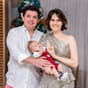 Software: Um casal sorridente está junto, segurando um bebê vestido de vermelho. Eles estão dentro de casa, com cortinas suavemente iluminadas e uma árvore de Natal decorada ao fundo, personificando a alegria do parto imediato nesta temporada de festas.