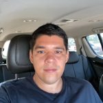 Software: Uma pessoa com cabelo curto e escuro está sentada no banco de trás de um carro, vestindo uma camiseta preta e olhando para a câmera. O interior de couro complementa a cena, e através da janela, outros veículos são visíveis, lembrando ícones em uma paisagem digital em uma loja de software.